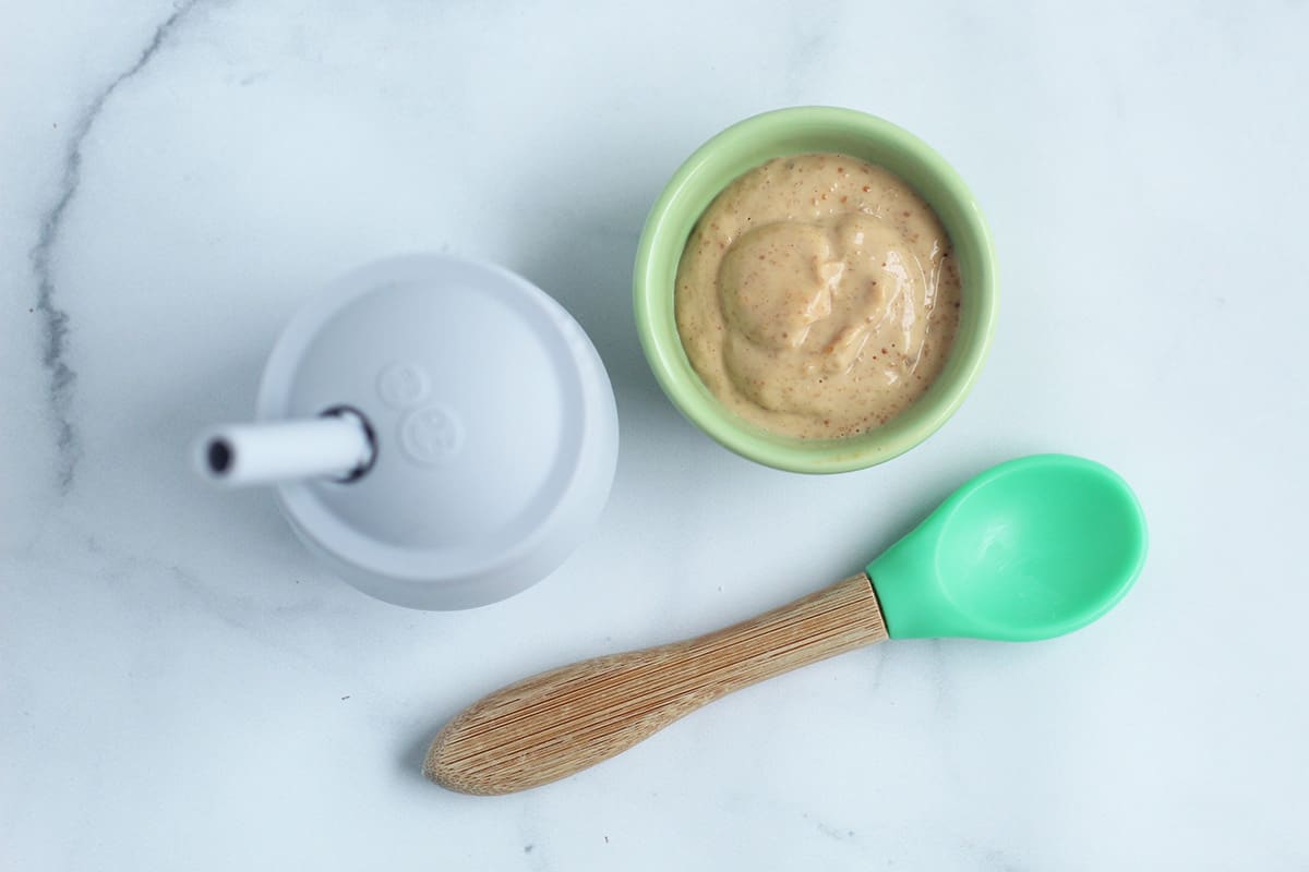 baby sippy cup with puree in bowl with green spoon on counter.