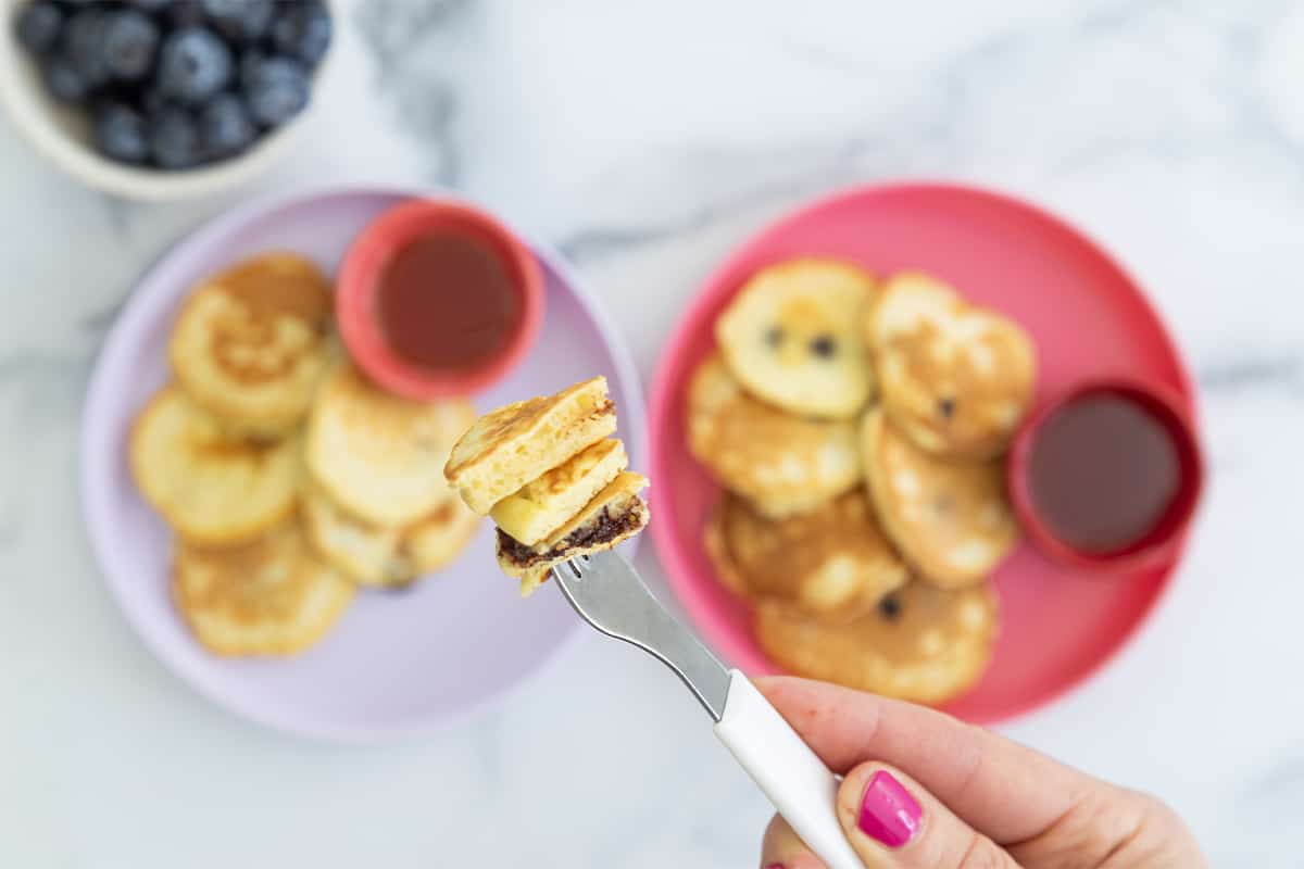 Chocolate chip pancakes served on kids plate with blueberries and syrup on the side and a bite ready on a fork.